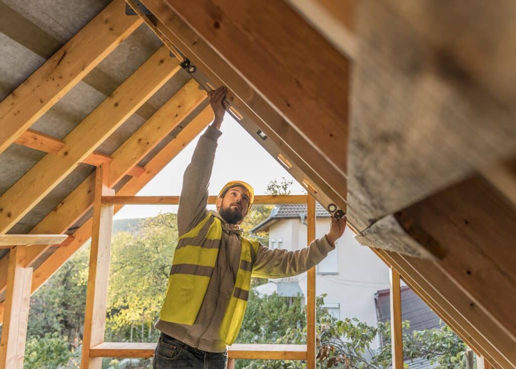 Charpentier en veste de signalisation et casque de sécurité installant ou vérifiant la structure en bois d'un toit en construction avec des maisons et des arbres en arrière-plan.
